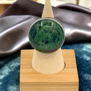 Op de afbeelding: Een ronde ring met een gepolijste groene en donker gespikkelde steen met een klein vleugje diepmagenta, gezet in een gladde zilverkleurige metalen rand. De ring wordt gepresenteerd op een beige kegelvormige standaard op een houten voet.