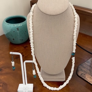 Op de afbeelding: Een witte schelpenketting en bijpassende oorbellen tentoongesteld op een sieraden buste en standaard. De ketting heeft turkooizen accenten. Op de achtergrond staat een turkooizen keramische pot.