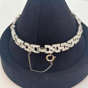 Könnte beinhalten: Ein silberfarbenes Metallarmband mit einem sich wiederholenden geometrischen Glieddesign, wobei jedes Glied mit kleinen klaren Strasssteinen besetzt ist. Das Armband verfügt über einen runden Federringverschluss und eine dünne Sicherheitskette.