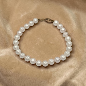 May include: A circular bracelet composed of uniform, round, white pearls strung together. The bracelet features a metallic, oval-shaped filigree clasp mechanism at the top, which connects the two ends of the pearl strand into a continuous loop.