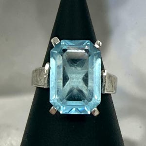 May include: A silver ring featuring a large, rectangular, emerald-cut light blue gemstone held in place by four prongs. The metal band has a textured, brushed finish on the sides, providing a contrast to the smooth, faceted surface of the stone.