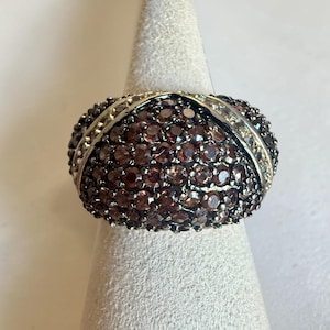 Pode incluir: Um anel de prata com uma parte superior abobadada, densamente coberto por pequenas pedras preciosas redondas, vermelho escuro. O anel tem uma faixa de prata com um padrão decorativo.