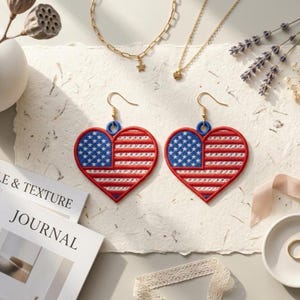 Op de afbeelding: Hartvormige oorbellen met een patriottisch ontwerp. De oorbellen tonen de Amerikaanse vlag, met een blauw veld met sterren en rood-witte strepen. Goudkleurige haken en blauwe accenten maken het ontwerp compleet. De oorbellen worden getoond met gouden kettingen en een tijdschrift.