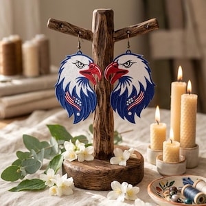 Può includere: Un paio di orecchini a forma di aquila su un supporto di legno. Gli orecchini presentano una testa bianca, becco rosso e dettagli blu e bianchi. L'esposizione include candele accese e fiori, creando una composizione decorativa.