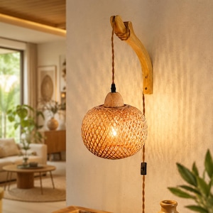 Op de afbeelding: Een wandlamp met een gebogen arm van lichtgekleurd hout en een bolvormige kap van gevlochten bamboe. De lamp hangt aan een gedraaid bruin stoffen snoer met een ingebouwde schakelaar en verspreidt een warm licht door het vlechtwerk.