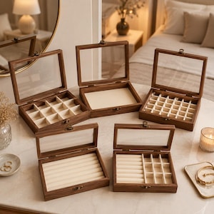Puede incluir: Cinco joyeros rectangulares de madera con tapas de cristal abatibles y cierres metálicos. Las cajas presentan diversas configuraciones interiores, incluyendo divisores de rejilla, rollos para anillos y compartimentos abiertos forrados con terciopelo color crema.