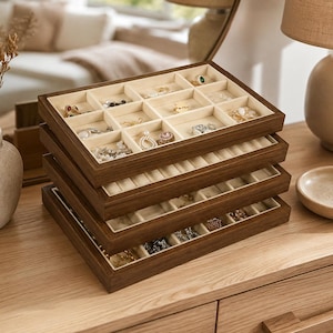 Peut inclure: Une pile de quatre plateaux organisateurs de bijoux rectangulaires en bois avec des cadres brun foncé et des intérieurs en velours crème. Chaque plateau comporte plusieurs compartiments pour ranger bagues, boucles d'oreilles et colliers.
