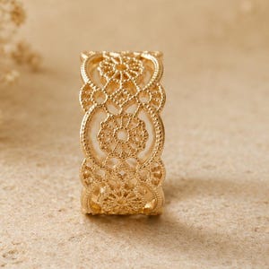 Puede incluir: Un anillo dorado con un intrincado diseño calado. El anillo presenta un patrón repetitivo de formas florales y geométricas, creando una apariencia delicada y ornamentada. El anillo se muestra sobre una superficie de tono neutro, resaltando sus detalles.