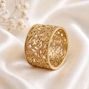Può includere: Un anello dorato con un intricato design traforato, caratterizzato da motivi floreali e a spirale. L'anello è largo e poggia su una superficie di raso bianco. Il design dell'anello è dettagliato e ornato, suggerendo uno stile vintage o antico.