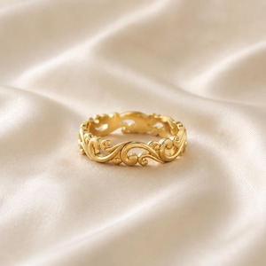 May include: A gold ring featuring an intricate, continuous band of raised, swirling scrollwork patterns. The metal has a polished, reflective finish that catches the light, highlighting the detailed, interconnected curves that form the circular structure.