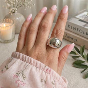 May include: A chunky, polished silver ring with a rounded, domed shape. The ring reflects the surroundings, creating a mirrored effect. The ring is worn on a hand with pink nail varnish, and a floral patterned sleeve is visible.