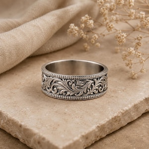 Può includere: Un anello in argento caratterizzato da un intricato motivo a volute floreali in rilievo lungo la fascia, bordato da due file di piccole perle metalliche in rilievo. Il metallo presenta una finitura lucida con aree incavate scure per risaltare il design.