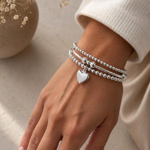 Può includere: Tre braccialetti d'argento su un polso. Due braccialetti sono con perline e uno è una fascia liscia. Un ciondolo a forma di cuore d'argento pende da un braccialetto. I braccialetti sono indossati su una mano con uno sfondo neutro.