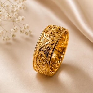 May include: A gold-colored ring with an intricate floral design. The ring is wide and has detailed carvings of flowers and leaves. The ring is set against a cream-colored satin background, with a sprig of white flowers in the upper left corner.