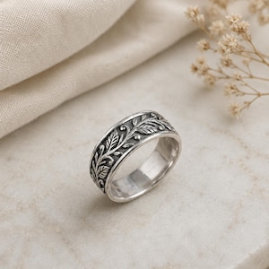 May include: A silver band ring featuring an intricate, raised relief pattern of leaves and vines wrapping around the exterior. The metal has a polished finish on the interior and a darkened, oxidised finish within the recessed leaf design.