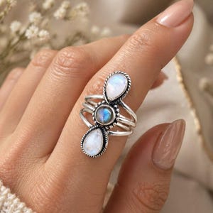 Puede incluir: Un anillo de plata con tres cabujones de piedra lunar. El anillo tiene una piedra central redonda y dos piedras en forma de pera, engastadas en una banda de plata detallada y texturizada. El anillo se lleva en un dedo.