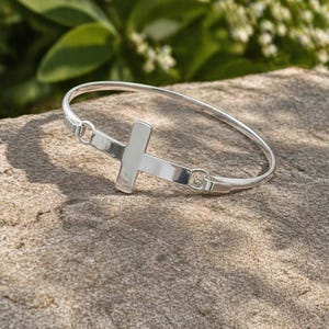 Puede incluir: Una pulsera rígida de tono plateado pulido que presenta un dije de cruz central. La cruz está integrada en la banda circular con dos pequeños aros de metal a cada lado, creando un diseño metálico continuo, elegante y reflectante.