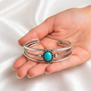 Puede incluir: Un brazalete de plata con una piedra turquesa ovalada central engastada en un bisel decorativo con textura de cuerda. La banda consta de tres hilos de plata paralelos, con el hilo central que presenta un diseño de cuerda trenzada y dos pequeñas esferas.