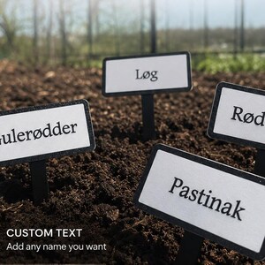 Pode incluir: Quatro marcadores de jardim retangulares brancos com texto preto são colocados em solo escuro. O texto nos marcadores diz: "Gulerødder", "Løg", "Rødbeder" e "Pastinak". O canto inferior esquerdo tem um ícone de lápis e o texto "CUSTOM TEXT Add any name you want".