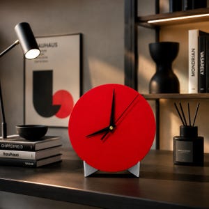 May include: A modern, minimalist red desk clock with black hands and a gold-colored center. The clock is round and sits on a white and silver stand. The clock is displayed on a dark surface with other decorative items.