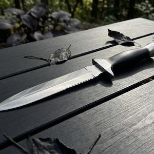 May include: A stainless steel dagger with a serrated edge and a black handle lies on a dark wooden surface. The knife is surrounded by fallen leaves, suggesting an outdoor setting. The blade is sharp and the handle is smooth.