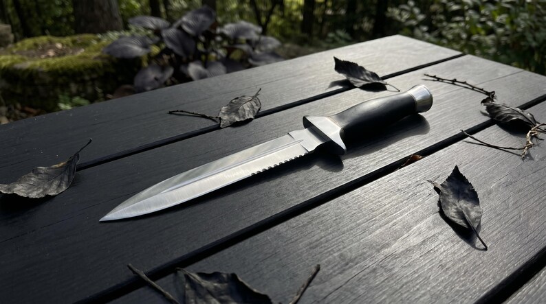 May include: A stainless steel dagger with a serrated edge and a black handle lies on a dark wooden surface. The knife is surrounded by fallen leaves, suggesting an outdoor setting. The blade is sharp and the handle is smooth.