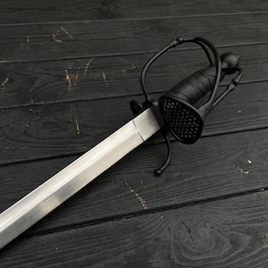 Könnte beinhalten: Ein schwarzes Rapier-Schwert mit silberner Klinge liegt auf einer dunklen Holzoberfläche. Der Griff hat einen komplexen Handschutz und einen strukturierten Griff. Die Klinge ist gerade und scharf und reflektiert das Licht.