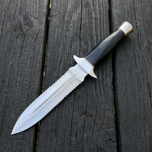 May include: A silver and black dagger with a serrated blade and a black handle. The knife has a silver guard and a silver and gold-coloured pommel. The knife is lying on a dark wooden surface.