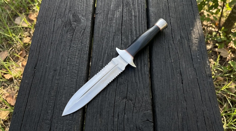 May include: A silver and black dagger with a serrated blade and a black handle. The knife has a silver guard and a silver and gold-coloured pommel. The knife is lying on a dark wooden surface.