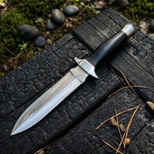 May include: A silver and black dagger with a serrated blade lies on a charred wooden surface. The handle is black with a silver guard and a gold-coloured end. The background includes moss, small stones, and pine needles.