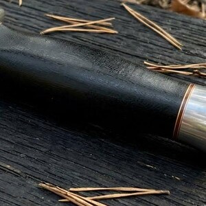 May include: A close-up of a knife handle featuring a black wooden grip and silver-coloured metal accents. The handle has a dark, textured appearance with a copper-coloured band near the metal. The knife rests on a dark wooden surface.