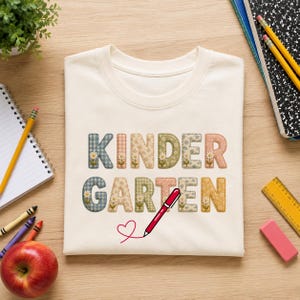 Puede incluir: Una camiseta de color crema con la palabra KINDERGARTEN en letras grandes y cuadradas con varios patrones florales y de cuadros. Un bolígrafo rojo aparece ilustrado debajo del texto, dibujando el contorno de un corazón rojo en la tela.