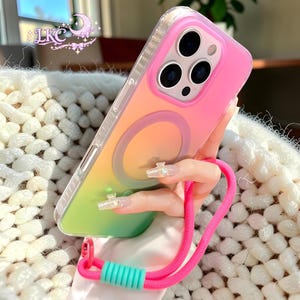 Puede incluir: Una funda para smartphone con un acabado degradado en rosa, naranja y verde, que presenta un anillo de carga magn&eacute;tico circular en la parte posterior. La funda incluye una correa de mu&ntilde;eca rosa brillante con una cuenta de ajuste turquesa y un borde protector transparente.