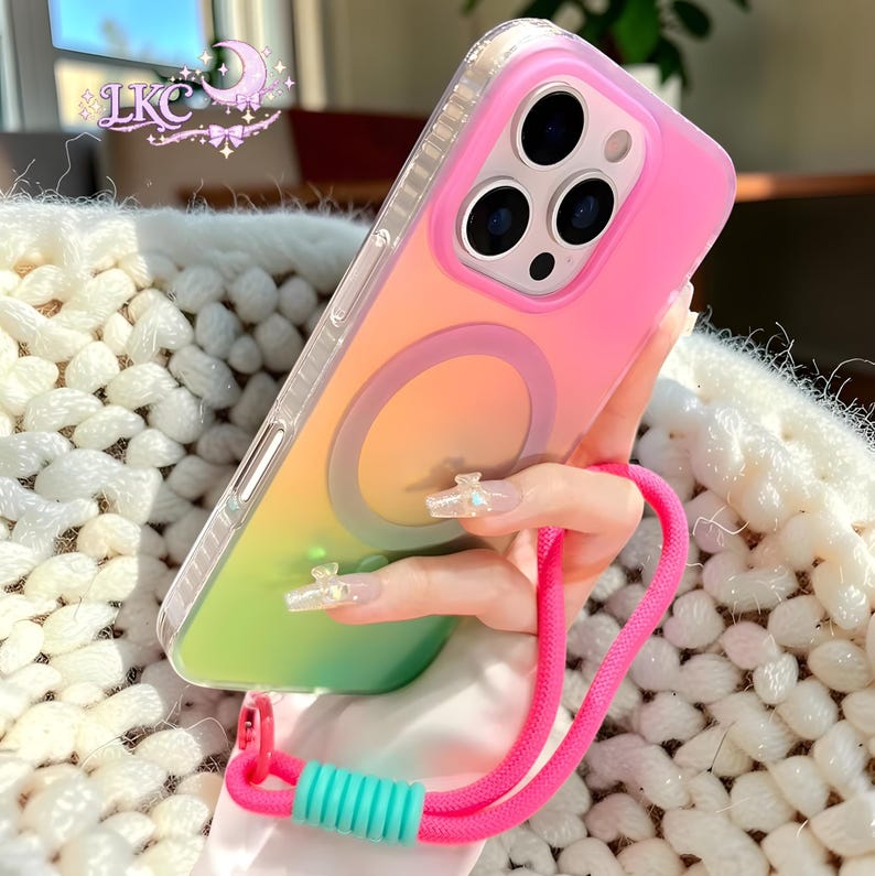 Puede incluir: Una funda para smartphone con un acabado degradado en rosa, naranja y verde, que presenta un anillo de carga magn&eacute;tico circular en la parte posterior. La funda incluye una correa de mu&ntilde;eca rosa brillante con una cuenta de ajuste turquesa y un borde protector transparente.