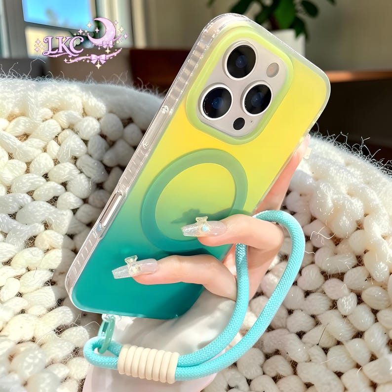 Puede incluir: Una funda para smartphone con un acabado degradado de amarillo a verde azulado y un anillo de carga magn&eacute;tico circular en la parte posterior. La funda incluye una correa de mu&ntilde;eca trenzada azul claro sujeta por un conector cil&iacute;ndrico color crema.