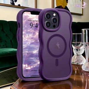Puede incluir: Una funda de smartphone de color morado con un diseño de bordes ondulados y festoneados, que incluye un anillo de carga magnético MagSafe circular en la parte posterior. La funda presenta un recorte elevado para la cámara y acabado mate.