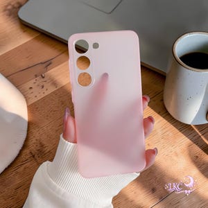 May include: A light pink phone case with cutouts for the camera and other features. The case is held in a hand with long nails. A laptop and a mug of coffee are in the background.