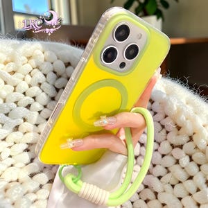 Puede incluir: Una funda de smartphone de color amarillo brillante con un borde transparente y un anillo de carga magn&eacute;tico circular en la parte posterior. La funda cuenta con una correa de mu&ntilde;eca verde claro en la parte inferior y un peque&ntilde;o logo en la esquina superior izquierda que dice LKC.