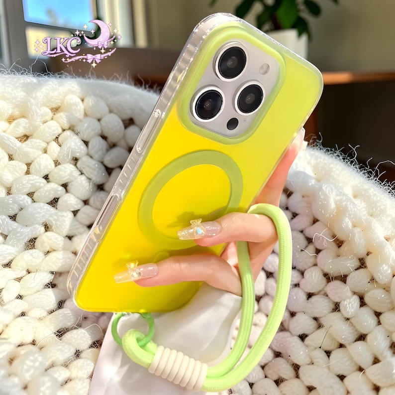 Puede incluir: Una funda de smartphone de color amarillo brillante con un borde transparente y un anillo de carga magn&eacute;tico circular en la parte posterior. La funda cuenta con una correa de mu&ntilde;eca verde claro en la parte inferior y un peque&ntilde;o logo en la esquina superior izquierda que dice LKC.