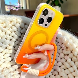 Puede incluir: Una funda de tel&eacute;fono con degradado amarillo y naranja brillante con un anillo de carga magn&eacute;tico circular en la parte posterior. La funda cuenta con un borde transparente y una correa de mu&ntilde;eca de cuerda naranja con conector blanco.