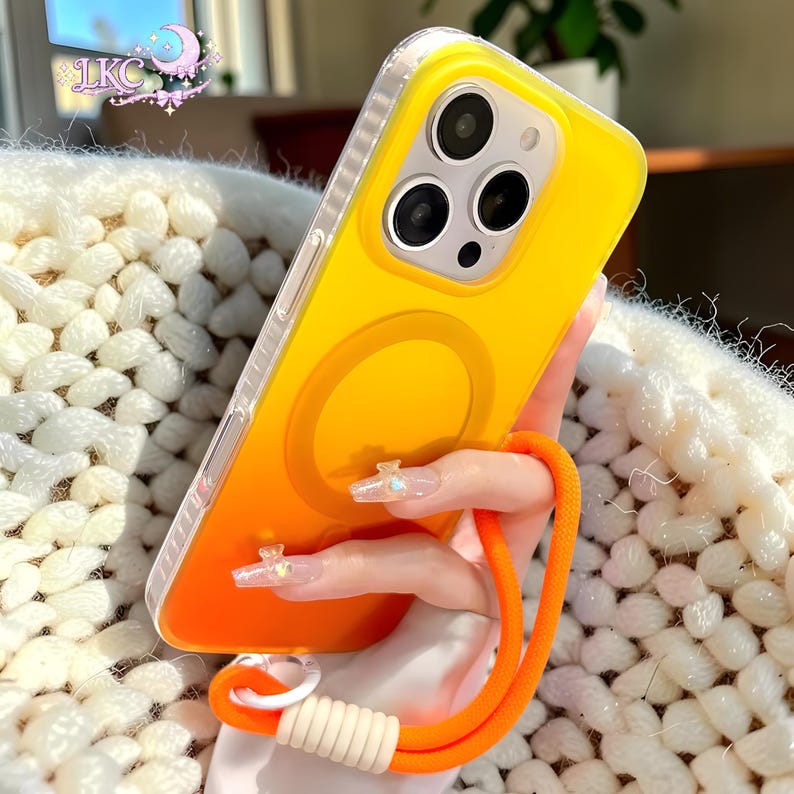 Puede incluir: Una funda de tel&eacute;fono con degradado amarillo y naranja brillante con un anillo de carga magn&eacute;tico circular en la parte posterior. La funda cuenta con un borde transparente y una correa de mu&ntilde;eca de cuerda naranja con conector blanco.
