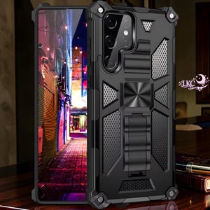 Puede incluir: Funda de teléfono negra con un diseño robusto. La funda tiene un soporte integrado y un panel trasero texturizado. La pantalla del teléfono muestra una colorida escena callejera. La funda tiene un acento metálico y esquinas protectoras.