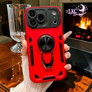 Puede incluir: Una funda de smartphone de color rojo brillante con una cubierta negra para la lente de la cámara, un soporte de anillo circular de metal negro en el centro y detalles negros en los laterales. Incluye un panel protector deslizante.