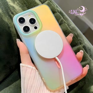 Puede incluir: Una funda para smartphone con un acabado de degradado suave que transiciona de azul claro a amarillo y rosa. La funda presenta un disco de carga magn&eacute;tico blanco circular en la parte posterior con un cable blanco hacia abajo. El texto LKC es visible.