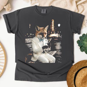 May include: A charcoal gray t-shirt featuring a graphic of a fox with a human body wearing a white tuxedo, a light blue bow tie, and black gloves, sitting on a bench while holding a martini glass with olives against a blurred city night background.