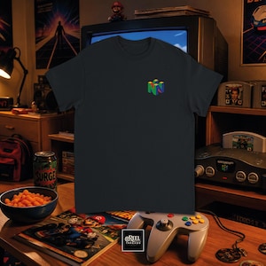 Puede incluir: Una camiseta de manga corta en color gris carbón oscuro que presenta un pequeño logotipo de Nintendo 64 multicolor estampado en la parte superior izquierda del pecho. La prenda tiene un cuello redondo clásico y un corte estándar.