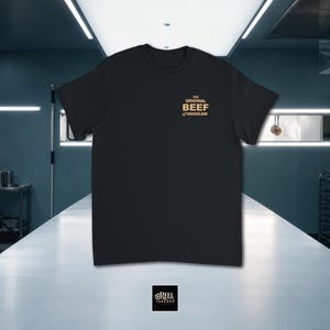 May include: A black short-sleeved t-shirt featuring a small tan graphic on the upper left chest area. The text reads THE ORIGINAL BEEF OF CHICAGOLAND in a stacked block font. The shirt has a crew neckline and a standard fit.