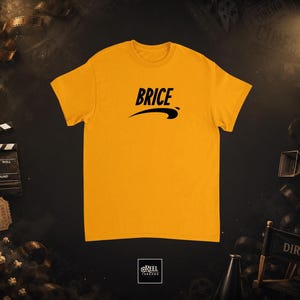 Peut inclure: T-shirt jaune moutarde avec le nom "BRICE" en noir, arqué au-dessus d'un trait noir. Le t-shirt est centré sur un fond sombre avec des accessoires de cinéma, dont un clap, une bobine de film et du pop-corn.
