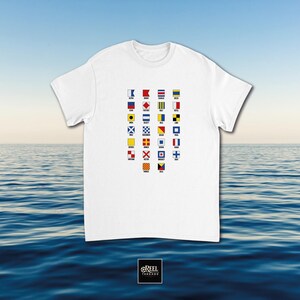 Op de afbeelding: Een wit t-shirt met korte mouwen en een centrale grafische print van het internationale maritieme seinvlaggenalfabet. Elke kleine vierkante vlag is in een raster geplaatst met de bijbehorende naam in zwarte letters eronder gedrukt.