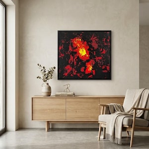 May include: A square framed wall art piece featuring an abstract composition with a black background and vibrant splashes of red, orange, and yellow paint. The artwork is displayed in a thin black frame, creating a bold contrast against the wall.